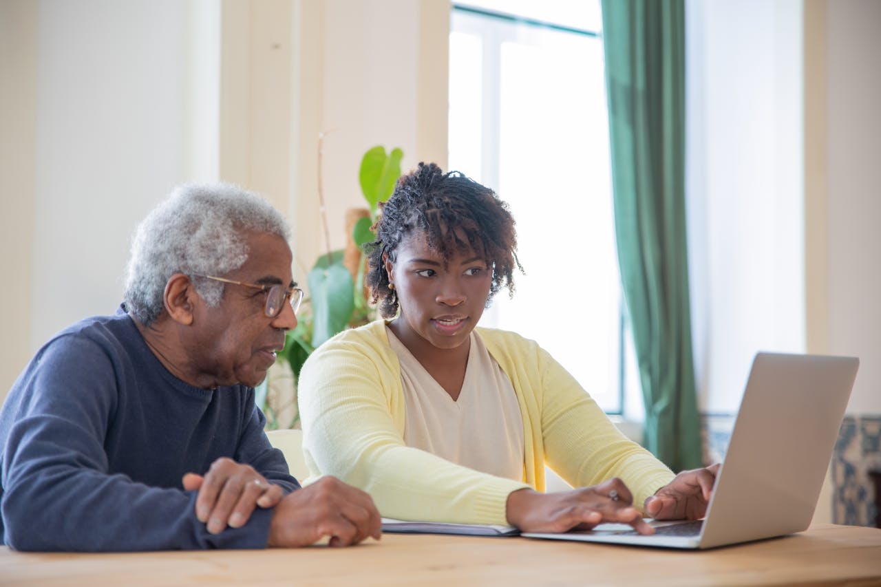 Young woman assists an elderly man with laptop indoors, highlighting intergenerational tech support.