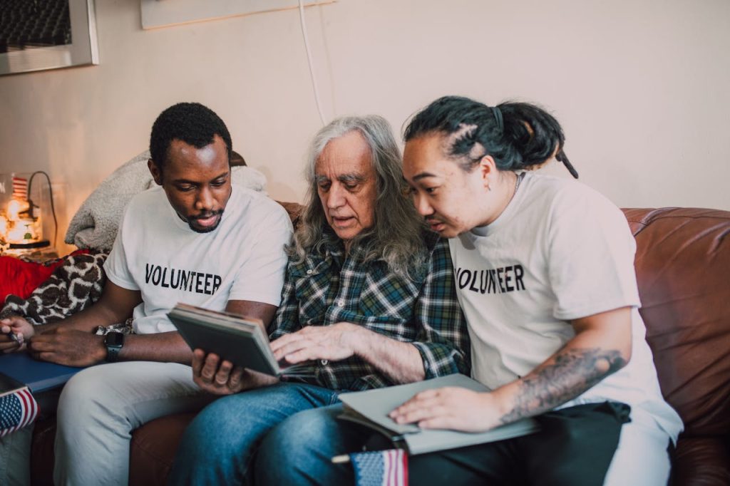 Volunteers helping an elderly man review a photo album during a home visit, fostering community support.